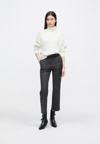 Λευκό πλεκτό πουλόβερ με ζιβάγκο, μαύρη ζώνη, γκρι κομμένα παντελόνια και μαύρες γόβες με τακούνι. Απλό φόντο, στάση όρθια.