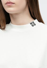 Biela bavlnená mikina s vysokým golierom a vyšívaným logom "HU GO" v čiernej farbe na ľavej strane goliera.