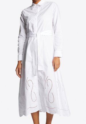 Femme portant une longue robe-chemise blanche avec col, ceinture à la taille, manches longues et motifs décoratifs ajourés près de l'ourlet.