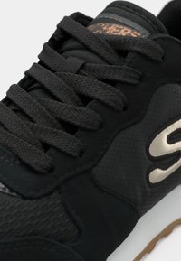 Close-up de um ténis Skechers preto mostrando o tecido texturizado, atacadores grossos e o logótipo da marca na lateral, com um detalhe em castanho visível na língua.