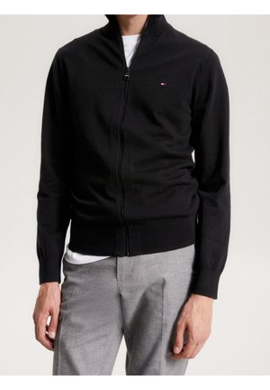 Homme portant un pull noir à fermeture éclair avec un petit logo sur la poitrine, un sous-vêtement blanc et un pantalon gris clair, debout devant un fond uni.