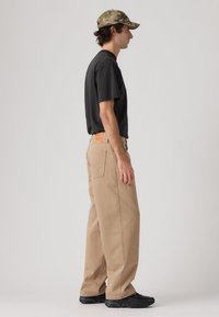 Μπεζ παντελόνια με φαρδιά πόδια συνδυασμένα με μαύρο μπλουζάκι και μαύρα αθλητικά παπούτσια. Το σύνολο ολοκληρώνεται με ένα καπέλο καμουφλάζ. Διακριτικές λεπτομέρειες στην πίσω τσέπη.