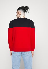 Červená a černá barevně blokovaná mikina s kulatým výstřihem, vyrobená z měkké tkaniny. Má dlouhé rukávy a žebrované manžety, v kombinaci se světle modrými džíny.