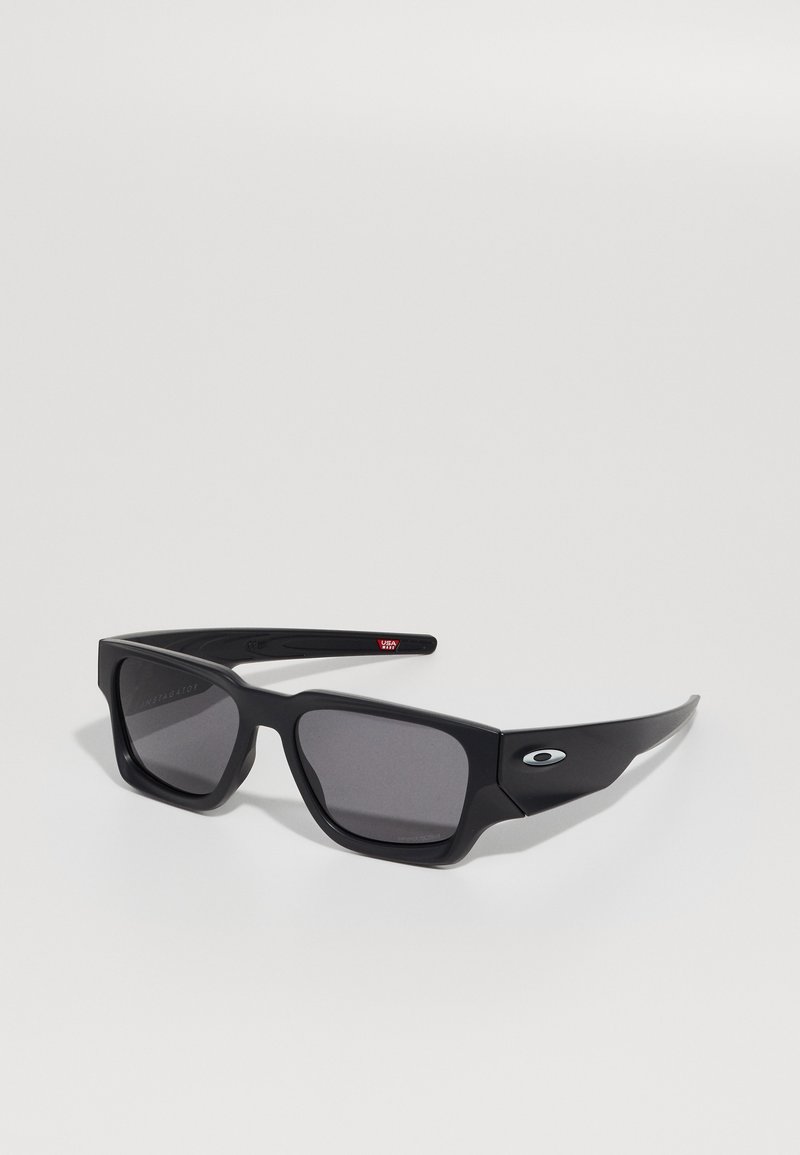 Gafas de sol rectangulares negras con lentes oscuros, montura gruesa y un logotipo en relieve en el costado. Presenta un acabado mate y un diseño robusto.