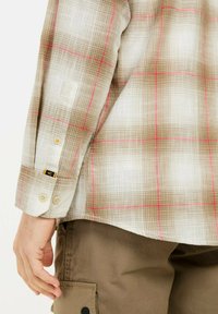 Lichte geruite shirt in tan en wit, met rode accenten, knoop manchetten en een gestructureerde stof. Gecombineerd met bruin cargobroeken.