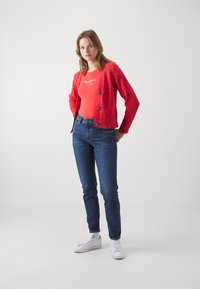 Červený pletený kardigan s textúrovaným vzorom a zapínaním na gombíky, kombinovaný s priliehavým červeným body a tmavomodrými džínami, nosený s bielymi teniskami.