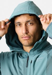 Türkisfarbene Kapuzenjacke mit strukturiertem Futter, Reißverschluss und Logo. Das Modell hält die Kapuze und präsentiert die Material- und Designdetails.