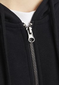 Sudadera con cremallera negra que presenta un cierre de cremallera plateado, tejido texturizado y una capa interior blanca visible en el cuello.