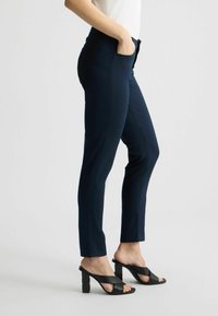 Pantalones de pinza de color azul marino, con un tejido suave, cintura alta, bolsillos laterales y bolsillos traseros; combinados con tacones negros con tiras cruzadas.