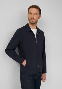 Marine zip-up jack met een vlakke kraag, lange mouwen, zijzakken en een gladde textuur, gecombineerd met een wit T-shirt en donkere jeans.