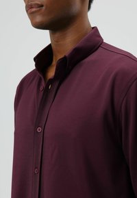 Bordeauxrood overhemd met lange mouwen en een gestructureerde kraag, voorzien van een gladde textuur en knoopsluiting aan de voorkant.