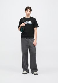 Μαύρο μπλουζάκι με λευκό λ ογότυπο, γκρι φόρμα και αθλητικά παπούτσια εκρού. Το μοντέλο στέκεται κρατώντας μια μικρή μαύρη τσάντα. Απλό φόντο.