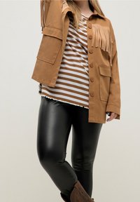 Giacca frangiata color tan con chiusura a bottoni, abbinata a una camicia a righe marrone e bianca, leggings neri lucidi e stivali alla caviglia marroni.