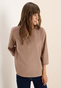 Femme aux cheveux bruns portant un haut beige ample et un jean bleu foncé, regardant vers la droite sur un fond clair et uni.
