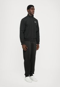 Black zip-up jacket with a stand collar, elastic cuffs, and side pockets. Worn over matching black pants and white sneakers. Simple design.