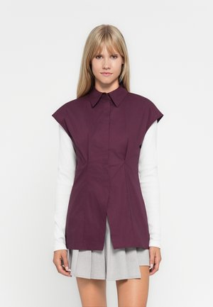 Camicia corta viola con colletto a punta e placket frontale, abbinata a una camicia bianca a maniche lunghe sotto e una gonna pieghettata grigia.