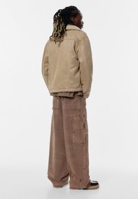 Jachetă bej căptușită cu blăniță sintetică, cu guler, asortată cu pantaloni maro cu croială largă, care au buzunare și un fit relaxat.