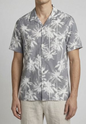 Man met een grijs kort shirt met witte palmbomenprint en lichtgekleurde broek tegen een effen achtergrond.