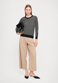 Pull en tricot noir et blanc à rayures avec des poignets côtelés, associé à un pantalon beige plissé et des chaussures noires à bout pointu. Sac à main noir.