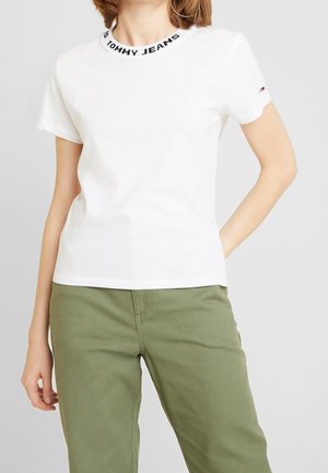 Personne portant un t-shirt blanc à manches courtes avec encolure et logo de manche de marque, associé à un pantalon vert olive, sur fond uni.