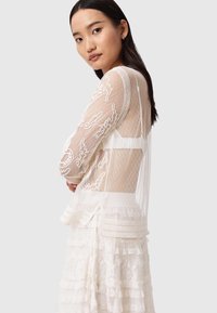 Transparente weiße Bluse mit langen Ärmeln, strukturierten Punkten und filigraner Stickerei. Mehrlagige Spitzenverzierung am Saum verleiht Tiefe.