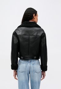 Γυναίκα που φοράει ένα μαύρο δερμάτινο μπουφάν �με γούνες στους γιακούς και τα μανίκια, συνδυασμένο με ανοιχτόχρωμες τζιν ψηλής μέσης, απεικονιζόμενη από πίσω.