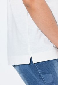 B�ílý top s krátkým rukávem a postranním rozparkem, vyrobený z měkké tkaniny, v kombinaci s modrými džíny, ukazující uvolněný střih a neformální styl.