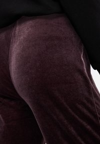 Μπορντό βελούδινες παντελόνες με μαλακή υφή, διαθέτοντας εφαρμοστή σιλουέτα και ελαφριά γυαλάδα. Μαύρο τοπ ορατό κατά την ανώτερη άκρη.