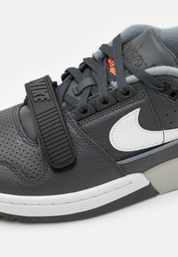 Nike tenisice s gornjim dijelom od sive kože, perforiranim dizajnom, bijelim logotipom i crnim remenom za sigurno prianjanje. Teksturirana potplat za bolju stabilnost.