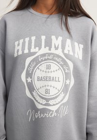 Šedá mikina so svetlými potlačami, vrátane nápisu "HILLMAN", dizajnu baseballovej lopty a "Norwich, UK". Mäkký materiál, voľný strih.