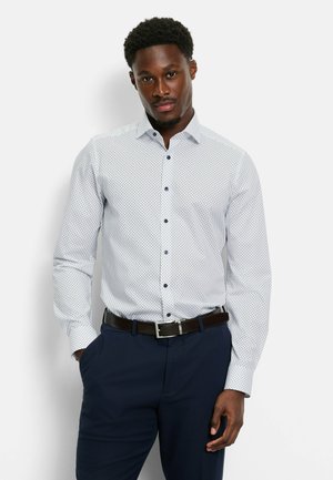 Witte blouse met kleine blauwe stippen, lange mouwen en een kraag. Gecombineerd met een donkere broek en een bruine riem.