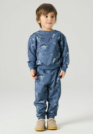Małe dziecko ubrane w niebieską bluzę i spodnie w dinozaury oraz beżowe buty, stojące na tle jasnej ściany.