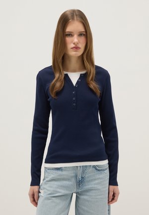 Jonge vrouw met lang lichtbruin haar, gekleed in een marineblauwe blouse met lange mouwen en knopen, gedragen over een wit ondershirt en lichtblauwe spijkerbroek.