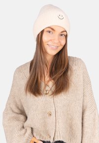 Bonnet en maille côtelée beige avec un emblème de visage souriant, porté avec un gilet beige épais à gros boutons dorés.
