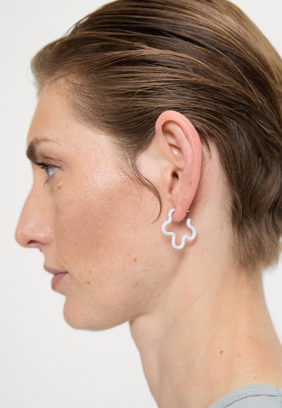 Bijele naušnice u obliku cvijeta s glatkom, sjajnom površinom, nošene na uhu manekenke. Dizajn je apstraktan s zakrivljenim linijama.