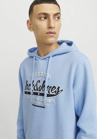 Svijetloplava hoodie izrađena od mekog materijala, s grafikom sprijeda u crno-bijelom tekstu, podesivom kapuljačom s vezicom i rebrastim manžetama.