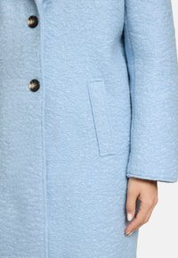 Manteau en laine bleu clair avec un tissu texturé, fermeture à boutons et poche latérale, silhouette structurée et manches larges.
