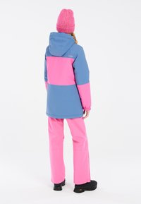 Chaqueta de nieve colorida en azul y rosa, con capucha, puños elásticos y tela duradera. Combinada con pantalones rosas a juego y botas negras.