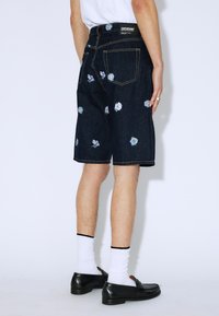 Pantalones cortos de denim oscuro con bordado floral, con un corte recto, bolsillos traseros y etiqueta negra en la cintura. Se llevan con calcetines y zapatos blancos.