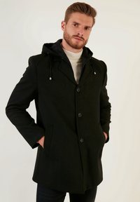 Abrigo negro con capucha, que presenta un cierre de botones en la parte delantera, bolsillos laterales y un diseño entallado. Tejido suave con una textura sutil.