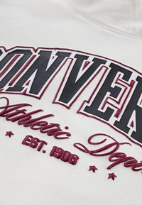 Sudadera blanca con texto grande en relieve. Las letras en negro y granate indican "CONVERSE Athletic Dept." con un "EST. 1908" estilizado abajo.