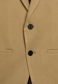 Marynarka khaki z fakturowanej tkaniny, jednorzędowa, z dwoma czarnymi guzikami. Posiada płaskie kieszenie oraz delikatne przeszycia jako detal.