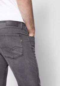 Vue arrière d'une personne portant un jean gris avec la main dans la poche avant et une chemise blanche sur un fond clair uni.