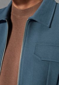 Blauwe gestructureerde jas met ritssluiting, voorzien van een kraag en een borstzak, gedragen over een bruine geribbelde trui.