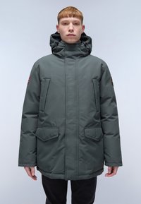 Giovane con capelli rossi corti che indossa un parka invernale verde scuro con cappuccio e diverse tasche frontali, in piedi su uno sfondo chiaro.