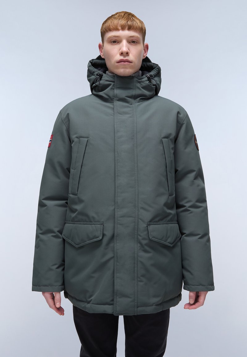 Jeune personne avec des cheveux roux courts portant un parka d'hiver vert foncé avec capuche et plusieurs poches à l'avant, se tenant devant un fond clair.