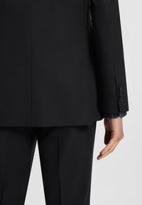 Completo nero su misura con una finitura testurizzata. La parte posteriore presenta una leggera apertura e polsini con bottone. Una camicia a fantasia è visibile sotto la giacca.
