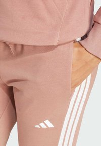 Růžové sportovní tepláky z měkkého materiálu s bílými pruhy po stranách a bílým logem. Obsahují boční kapsu.