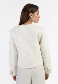 Blazer bianco texturizzato con spalle imbottite, scollatura rotonda e bottoni dorati sulle maniche, abbinato a pantaloni beige.