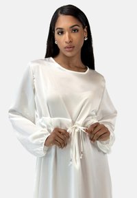 Witte satijnen jurk met lange, opgepofte mouwen, elastische manchetten en een taille met trekkoord. Heeft een gladde textuur en een subtiele glans.
